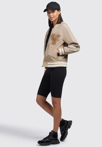 Beige bomberjack met geborduurd tijgerontwerp, witte strepen op ribgebreide manchetten, gecombineerd met zwarte fietsbroek en zwarte sneakers.