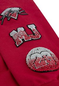 Tessuto rosso con tre elementi ricamati: "Magia" in bianco e nero, "MU" in grigio testurizzato e un'applicazione circolare con "23" in grigio e rosso.