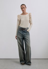 Donna che indossa un top lungo beige con texture e spalle a sbuffo, jeans a gamba larga sbiaditi, cintura doppia con borchie e scarpe a punta.
