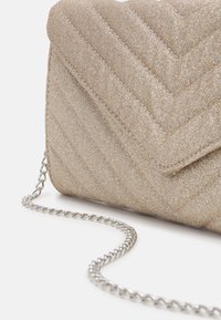 Błyszcząca beżowa pikowana torebka kopertowa na łańcuszku. Posiada wzór jodełki, gładką fakturę i spiczasty klapkę z zapięciem.