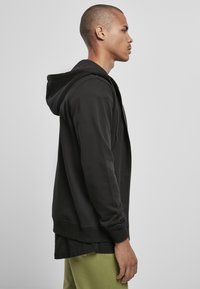 Sudadera negra con cremallera y textura suave, con bolsillo tipo canguro, llevada sobre una camiseta oscura, mostrada de lado sobre un fondo liso.