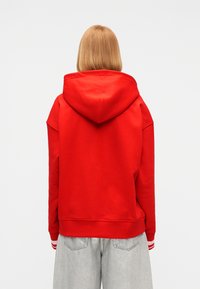 Άτομο με ίσια, μακριά μέχρι τους ώμους μαλλιά φορώντας φωτεινό κόκκινο φούτερ με κουκούλα και ανοιχτόγκρι τζιν, γυρισμένο προς τα πίσω σε απλό φόντο.