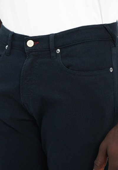 Pantalones de mezclilla de un azul oscuro que presentan un diseño clásico de cinco bolsillos, cierre de botón metálico y cinturas cosidas, con una textura sutil.