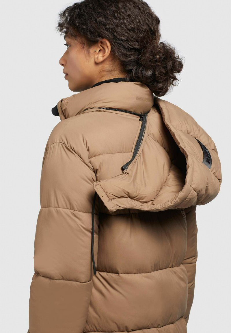 Eine Person mit lockigem Haar, das nach hinten gebunden ist, trägt eine beige Steppjacke mit einer teilweise offenstehenden, hängenden Kapuze und steht nach links vor einem schlichten Hintergrund.