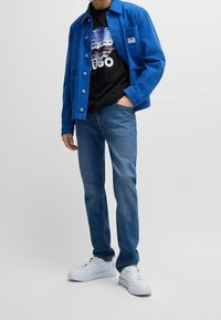 Blå jacka, svart grafisk t-shirt med palmönster, blå jeans och vita sneakers. Jackan har en bröstficka och knappstängning.