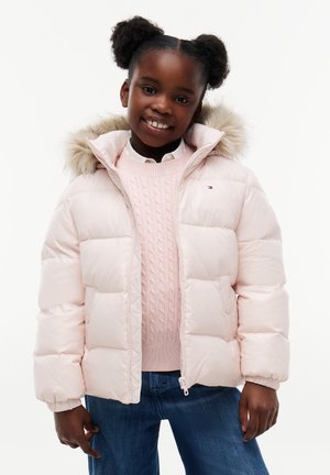 Lächelndes Mädchen mit zwei Puff-Zöpfen trägt eine hellrosa gesteppte Jacke mit Fellkapuze, einen rosa Zopfstrickpullover und blaue Jeans vor weißem Hintergrund.