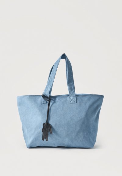 Sac cabas en toile bleu clair avec deux anses, un petit logo brodé et un porte-bonheur noir en forme de crocodile, sur un fond uni.