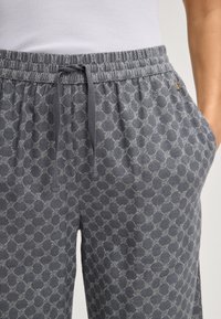 Šedé vzorované tepláky s elastickým pasem, tkaničkou a bočními kapsami. Tkanina má geometrický design ve světlé šedi.