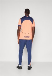 Sportoutfit med en koralfarvet kortærmet top med marineblå detaljer og 'UNHCR' branding, parret med marineblå joggers og sorte sneakers.