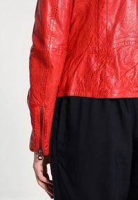 Czerwona skórzana kurtka o dopasowanym kroju; posiada fakturowany materiał, widoczne przeszycia oraz suwak na rękawie.
