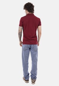 Uomo con capelli ricci e braccio sinistro tatuato, indossa una polo bordeaux, jeans azzurri e sneakers Nike marroni, visto di schiena.