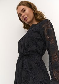 Svart spetsklänning med långa ärmar, intrikata blommönster, ett knytbälte i midjan och en texturerad finish. Strukturerad krage och knappdetaljer.