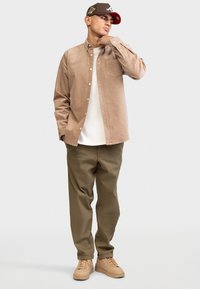 Beige Corduroy Hemd mit Knöpfen, darunter ein weißes T-Shirt, olivgrüne Hose und hellbraune Sneakers; trägt eine braun-rote Mütze.