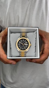 Montre-bracelet ton argent et or dans une boîte, avec un cadran noir, un affichage de la date et un bracelet en acier inoxydable. Enveloppée dans un tissu gris doux.