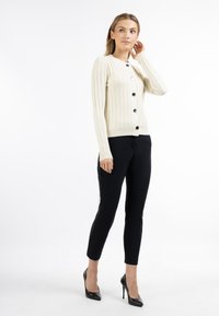 Cardigan a coste crema con bottoni neri, abbinato a pantaloni neri a sigaretta e tacchi neri, su uno sfondo neutro.