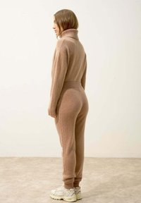 Beżowy dzianinowy kombinezon z wysokim kołnierzem, prążkowanymi mankietami i zwężanymi nogawkami. Posiada detale w stylu kablowym wzdłuż boków.