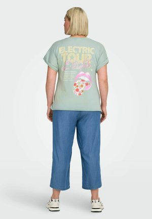 Kvinde med kort blondt hår iført en lysegrøn T-shirt med teksten "Electric Tour" og et blomstermotiv på tungen, blå bukser og sneakers.