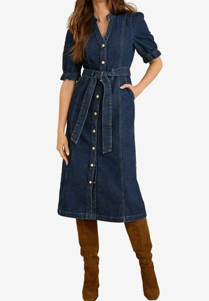 Kvinde iført en mørkeblå denim midi-kjole med knapper foran, bælte i taljen, pufærmer og brune ruskind-knæstøvler.