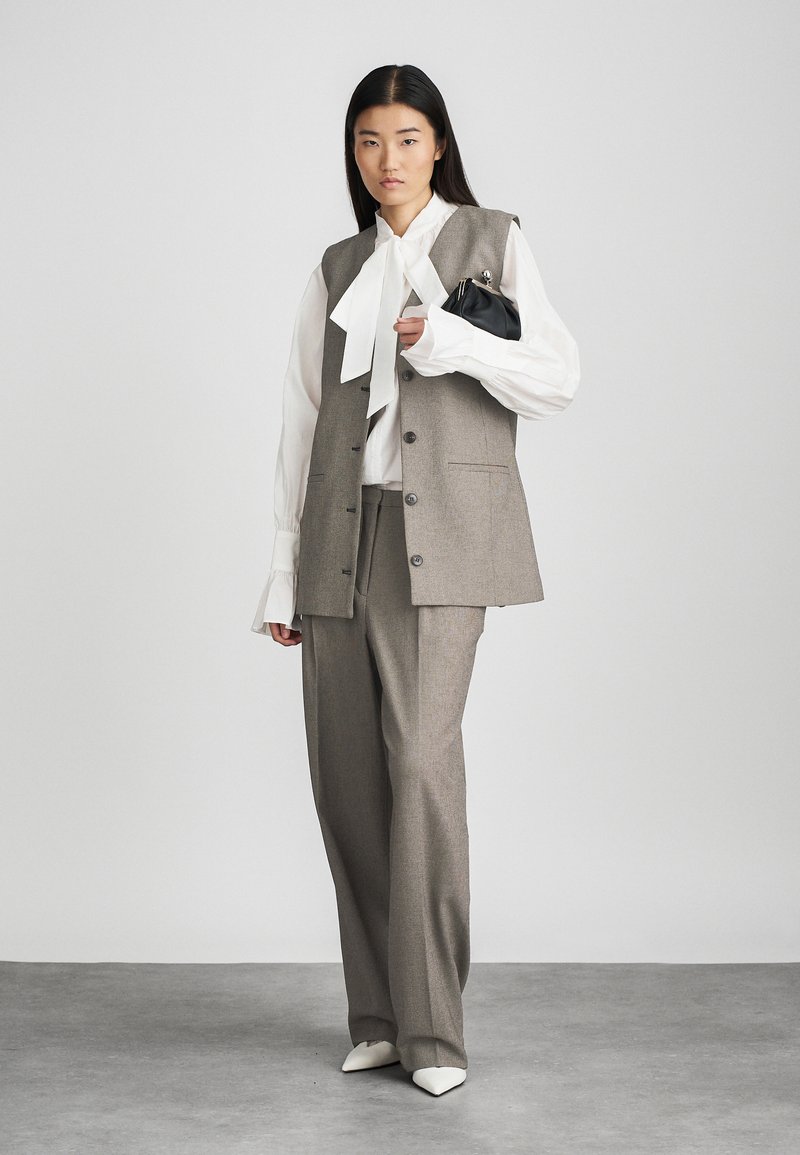 Grijze op maat gemaakte gilet met knopen, gecombineerd met hoge grijze broek. Witte blouse met een grote strik. Accessoires: kleine zwarte handtas.