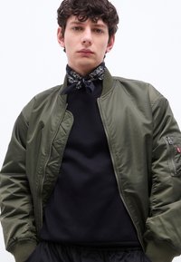 Νέος άνδρας που φοράει πράσινο μπουφάν bomber πάνω από μαύρο πουκάμισο, διακοσμημένος με μαύρο φουλάρι με σχέδιο, στέκεται μπροστά σε λευκό φόντο.