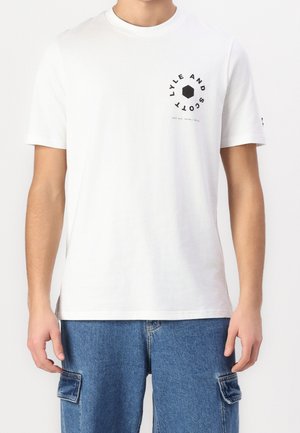 Mand iført hvid T-shirt med korte ærmer med sort cirkulært "LYLE AND SCOTT" logo på brystet og blå denim cargo shorts.