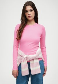 Dugih rukava roza rebrasti top s okruglim izrezom, uparen s bijelim i roza prugastim pletenim puloverom zavezanom u struku. Plave traperice.