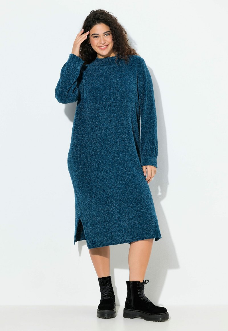 Abito in maglia a maniche lunghe di colore teal, realizzato in tessuto a maglia testurizzato, con spacchi laterali per facilitare i movimenti, abbinato a stivali neri con lacci alla caviglia.