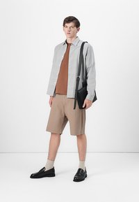 Ung mann iført grå rutete overskjorte, brun t-skjorte, beige stripete shorts, svarte loafers, som holder en svart skulderveske på hvit bakgrunn.