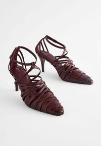 Sandalias de tacón alto burdeos con punta afilada, que presentan intrincadas correas entrelazadas y un acabado suave y brillante. Hebilla ajustable en el tobillo.