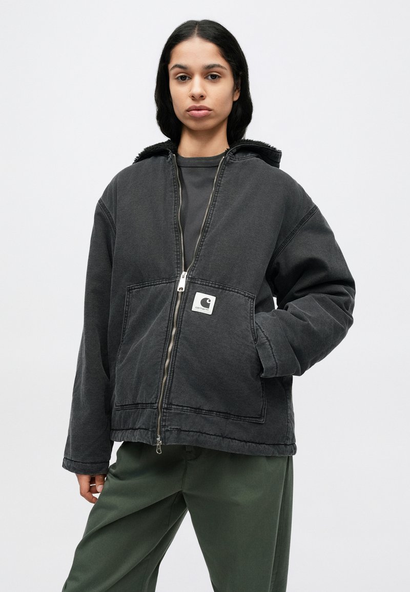 Schwarze Jeansjacke mit Reißverschluss vorne, großen Taschen und einem Kapuze mit Kunstfellfutter. Mit einem Logo-Patch auf der linken Brust. Kombiniert mit grünen Hosen.