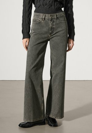 Hoch taillierte, ausgewaschene schwarze Jeans mit weitem Bein, kombiniert mit einem schwarzen Zopfpullover und schwarzen Lacklederloafern vor einem neutralen Bodenhintergrund.