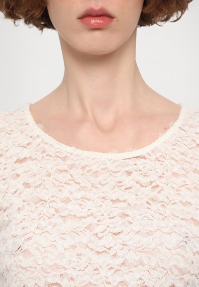 Krajková blůza v jemně bílé barvě, s květinovým vzorem, kulatým výstřihem a jemně vykrojenými okraji. Průhledná textura s decentním pokrytím.