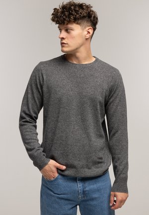 Szary sweter dziany z okrągłym dekoltem, długimi rękawami i prążkowanymi mankietami, w zestawie z jasnoniebieskimi dżinsami. Posiada fakturowaną tkaninę oraz subtelny detal z logo.