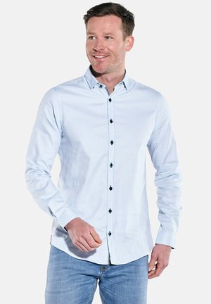 Man wearing light blue buttoned shirt and jeans, smiling and looking slightly right, standing against plain white background.