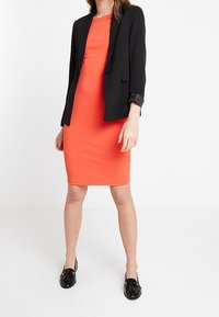 Svart blazer över en orange figurnära klänning, knälång. Släta tyger, enfärgade, svarta slip-on-skor med en blank yta.