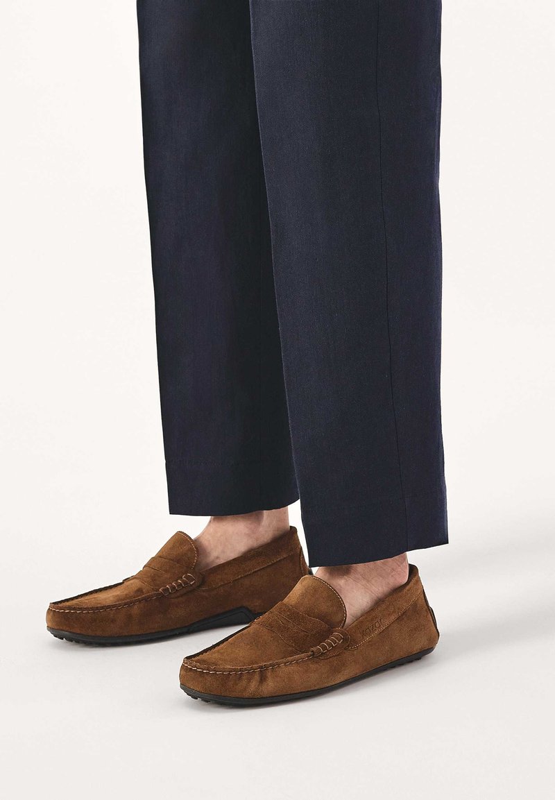 Mocassini in camoscio marrone indossati con pantaloni blu navy corti, mostrati su sfondo chiaro.