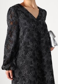 Black dress with floral embroidery, V-neckline, puffed sleeves, and sheer texture. Silver clutch held in right hand.