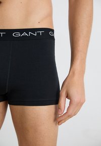Mandlig torso og øvre lår iført sorte boxershorts med GANT-logo på linningen, stående med højre arm afslappet langs siden.