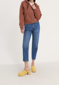 Sudadera marrón corta con cremallera, vaqueros azules de pierna recta y zuecos amarillos con tacón. Tela suave, ajuste informal y textura lisa.