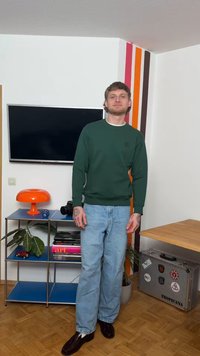 Νέος άνδρας με τατουάζ, φορώντας πράσινο φούτερ και τζιν, στέκεται σε δωμάτιο με τηλεόραση, πορτοκαλί λάμπα, ράφια και ριγέ τοιχογραφία.