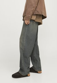 Person wearing loose-fit gray jeans, brown layered tops, and black slip-on shoes standing against a plain light background.