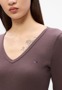 Top maro cu guler în V, cu dungi, având un mic logo brodat în albastru, alb și roșu pe partea stângă, cu o textură de țesătură netedă.