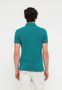 Πουκάμισο πόλο τυρκουάζ με γιακά και κοντά μανίκια. Κατασκευασμένο από υφασμάτινη ύφανση, με ίσιωμα στη βάση και εφαρμοστή γραμμή, συνδυασμένο με ανοιχτόχρωμο παντελόνι.