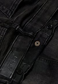 Svart denimstoff med en lett slitt tekstur, med et lærmerke, svarte knappefester og en forsterket midje.