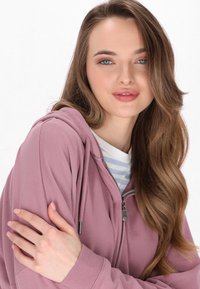 Roze zip-up hoodie met een gestructureerde pasvorm, trekkoordcapuchon en zilveren rits; gedragen over een wit gestreept shirt. Gladde textuur.