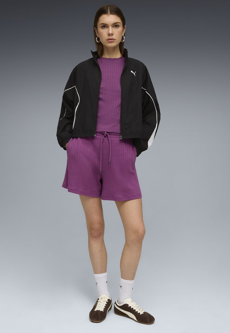 Donna in piedi con le mani nelle tasche, indossa una giacca nera Puma, pantaloncini e top viola a coste, calzini bianchi e scarpe da ginnastica scure su sfondo grigio.