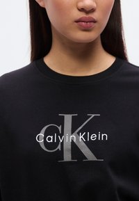 Μαύρο βαμβακερό φούτερ με μεγάλο γκρι λογότυπο "CK" και λευκό λογότυπο "Calvin Klein". Κλασικό λαιμόκοψη crew με απαλή υφή.