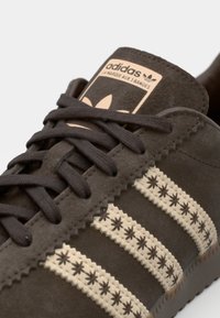 Sneaker in camoscio marrone con tre strisce zigzag beige, dettagli testurizzati e un'etichetta beiga sulla linguetta con il logo Adidas.