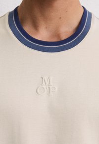 Primo piano di una camicia beige con colletto a righe blu e bianche e logo ricamato "M O’P" sollevato sul petto.