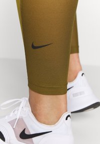 Olive zielone legginsy Nike z teksturowanej tkaniny mają czarne logo Nike. W zestawie z białymi butami sportowymi z przezroczystymi panelami i czarnymi akcentami.